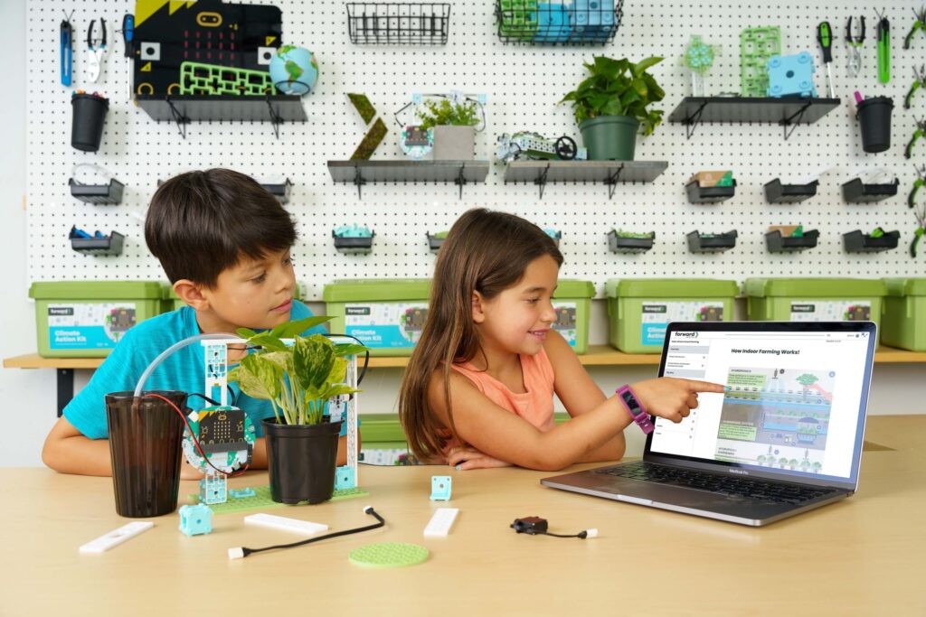 Two children engaged in a hands-on STEAM activity in the Forward Education workshop space. The smart farming project featuring a micro:bit and Climate Action Kit components is set up on the desk. One child points to a laptop screen displaying an educational diagram titled "How Indoor Farming Works."