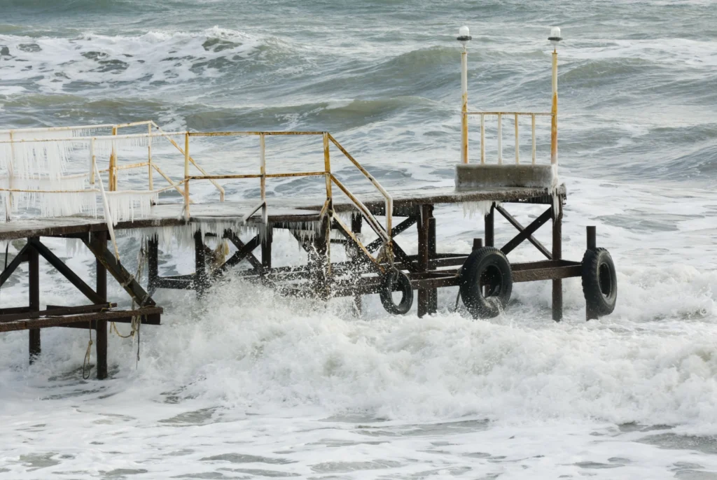 frozen pier with icicles hanging from its structure as waves crash below in a rough sea.