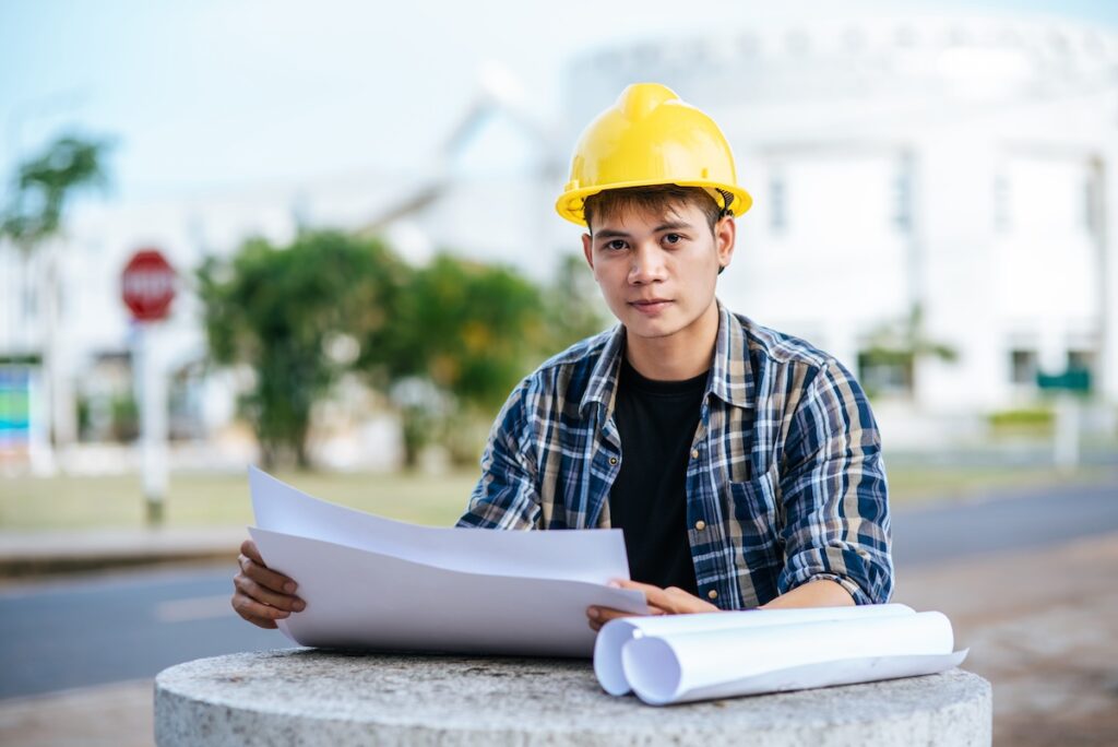 A man looking into the camera wearing a hardhat while holding rolls of paper.