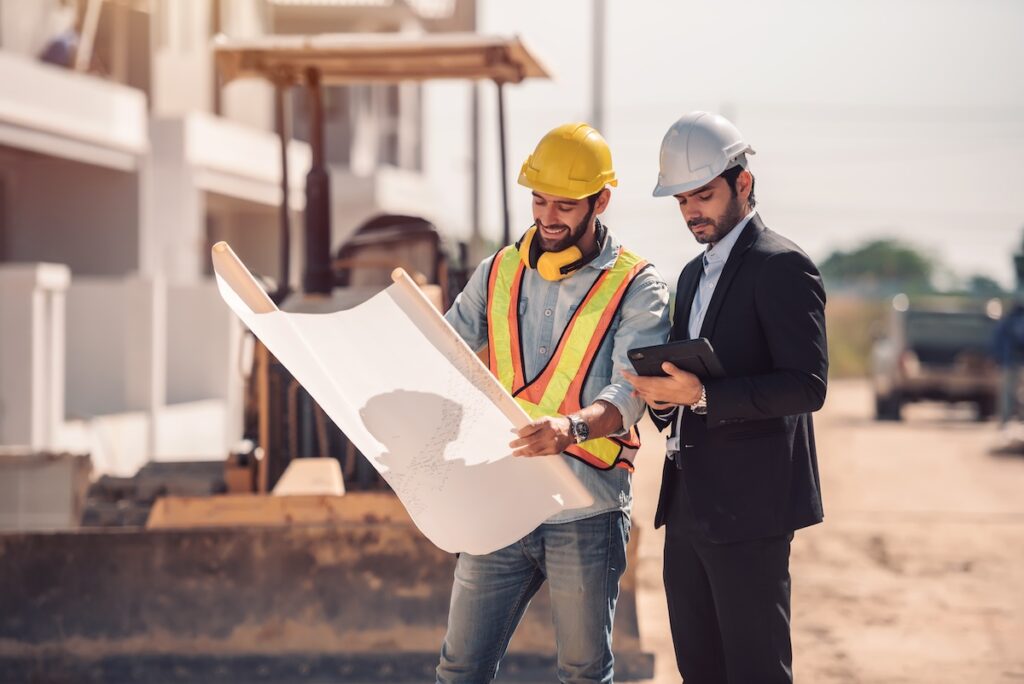 Two people in hard hats on a construction site looking at a large roll of paper.