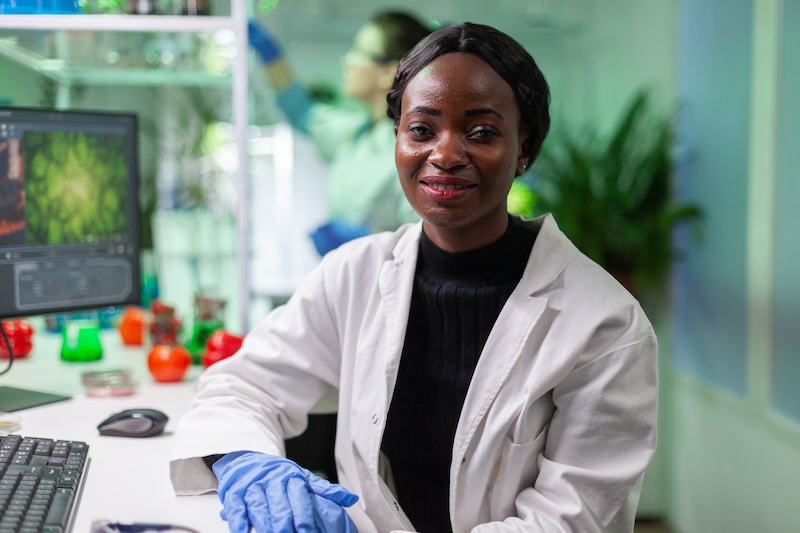 Closeup of african biologist woman looking into camera while working in biological agronomy laboratory. Specialists team researching genetic mutation developing medical scientific GMO dna test.