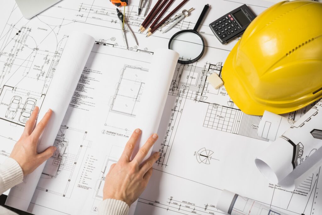 Hands on a table with a hardhat and blueprints for a building.
