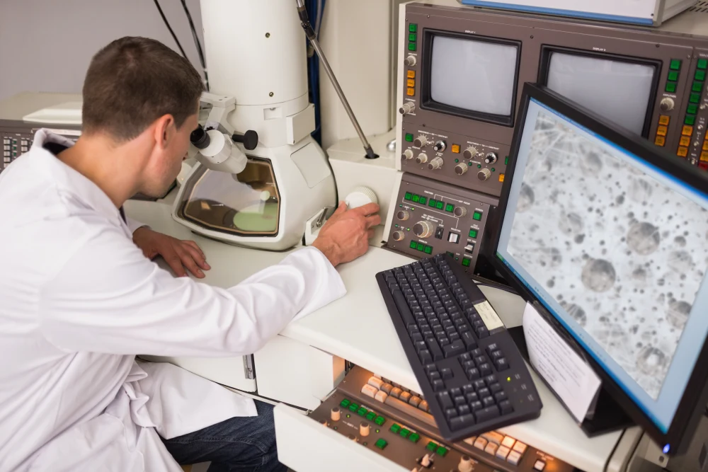 A scientist looking through a large desktop microscope. A monitor beside him is displaying the results.
