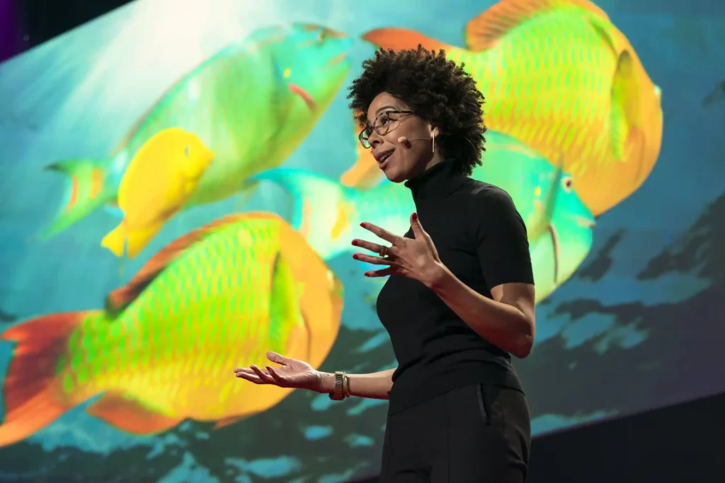Ayana Elizabeth Johnson giving a speech in front a digital screen of yellow swimming fish at a TedTalk