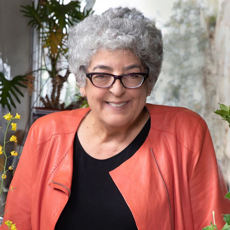 Joanne Chory an older lady with a grey perm stands for a portrait surrounded by plants in a science lab.