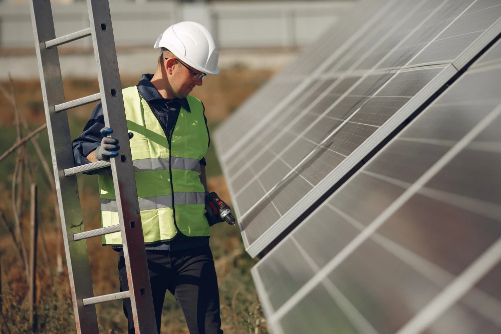 A technician inspecting a solar panel. He is also carrying a ladder and a drill.