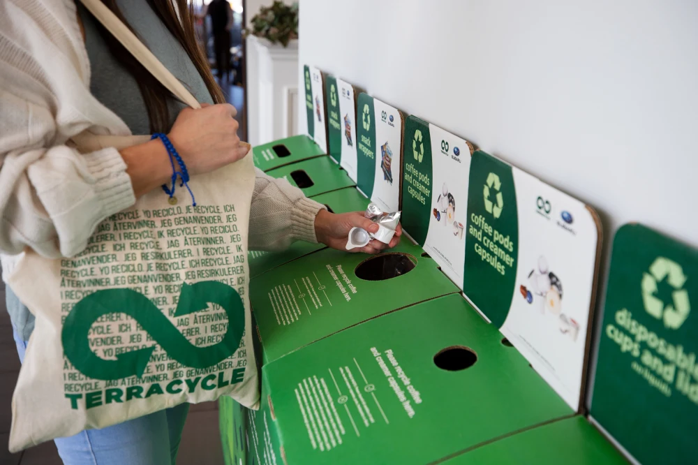 A row of TerraCycle recycling bins along a wall. A person carrying a TerraCycle tote bag standing in front of the bins placing plastic into one of the bins.