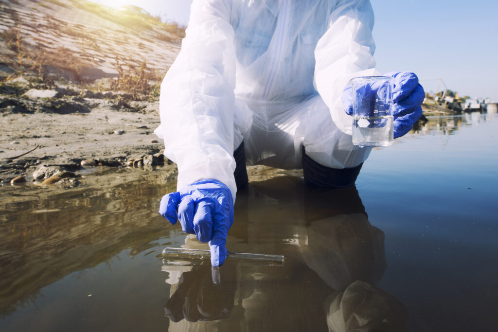 A scientist wearing protective gloves and a white suit collects water samples. The image illustrates environmental monitoring and efforts to measure pollution levels in a body of water.
