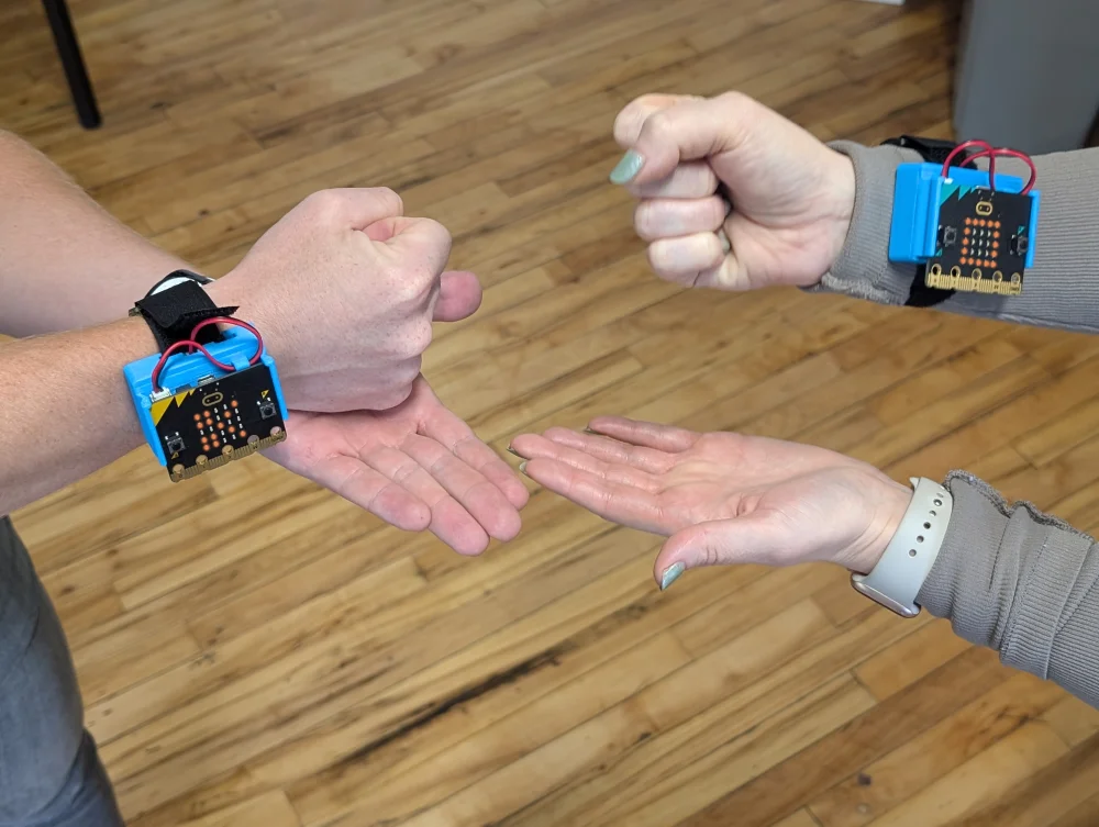 Two people's hands playing Rock Paper Scissors with CHARGE power packs and micro:bits attached to their wrists.