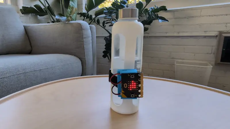 Demonstration of water bottle with the CHARGE for micro:bit attached, the micor:bit is displaying the duck icon and then being lifted out of frame, then set back onto the table with an X on the micro:bit. Then resetting the micro:bit to display the duck icon.