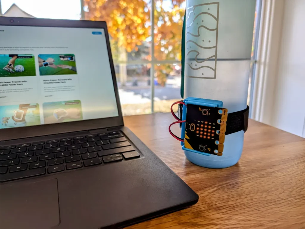 A water bottle with a CHARGE power pack and micro:bit attached to it sits on a table beside a laptop.