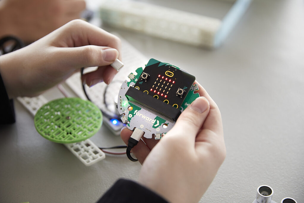 A close-up of hands holding a BBC micro:bit attached to the Climate Action Kit's breakout board. The board has several connected wires and components, part of a project setup. Other Climate Action Kit building blocks are visible on the table in the background, indicating a work-in-progress electronics or robotics activity.