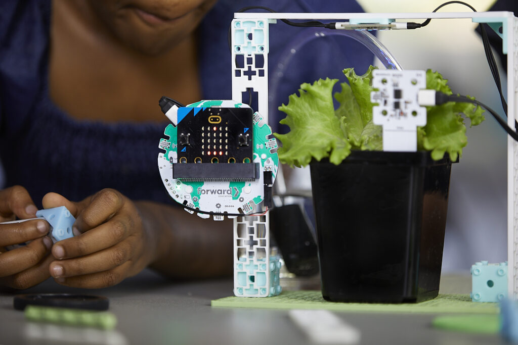 A close-up of a hands-on project involving a BBC micro:bit integrated into a hydroponic system built with the Climate Action Kit. The micro:bit is mounted onto the breakout board, and attached to a white structural frame. A black potted plant with green lettuce is connected to the system, suggesting an automated plant monitoring or irrigation setup. A person in the background is interacting with components, assembling or adjusting the system.