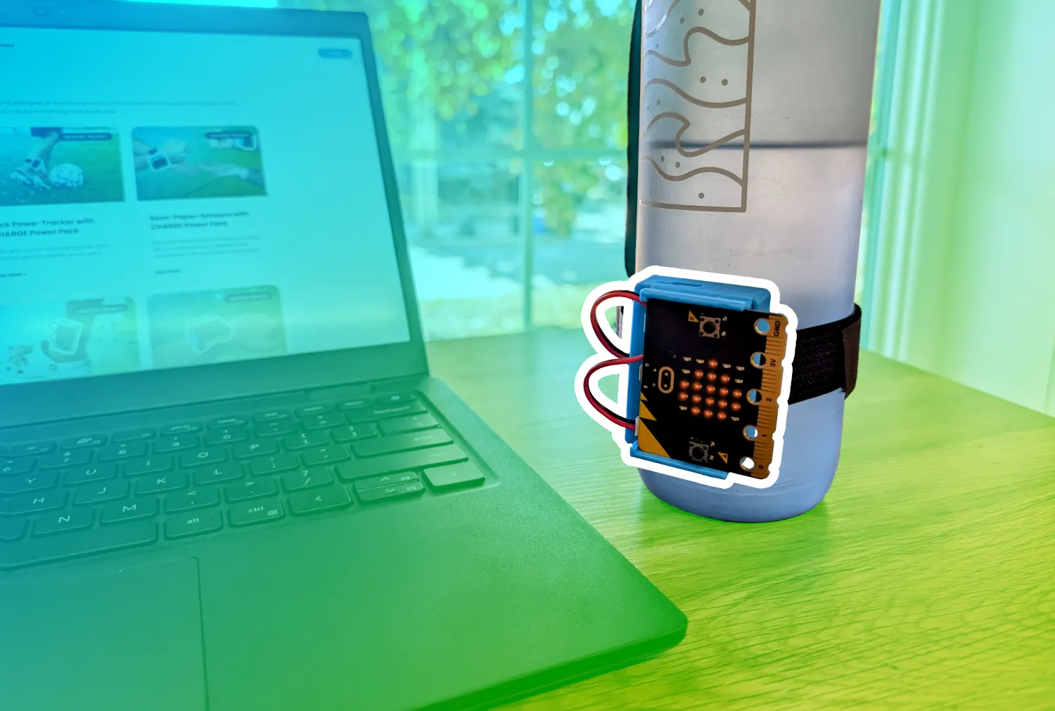 A water bottle with a CHARGE power pack and micro:bit attached to it sits on a table beside a laptop. A blue to green gradient is overlaid behind the micro:bit which has a white boarder around it.