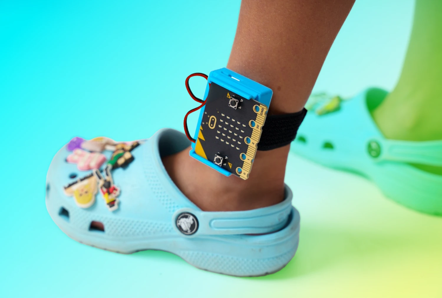 Closeup of a Charge for micro:bit wrapped around a child's ankle wearing crocs. A blue to green gradient is overlaid behind the micro:bit which has a white boarder around it.