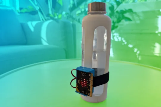 Water bottle sitting on a table with a CHARGE for micro:bit attached to the front of it. A blue to green gradient is overlaid behind the micro:bit which has a white boarder around it.