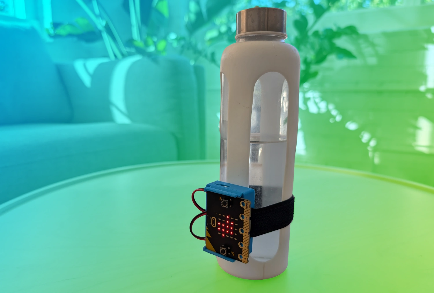 Water bottle sitting on a table with a CHARGE for micro:bit attached to the front of it. A blue to green gradient is overlaid behind the micro:bit which has a white boarder around it.