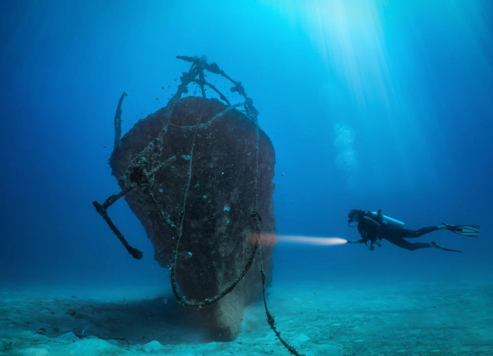 A solo diver with a flashlight swims toward the bow of an upright shipwreck.