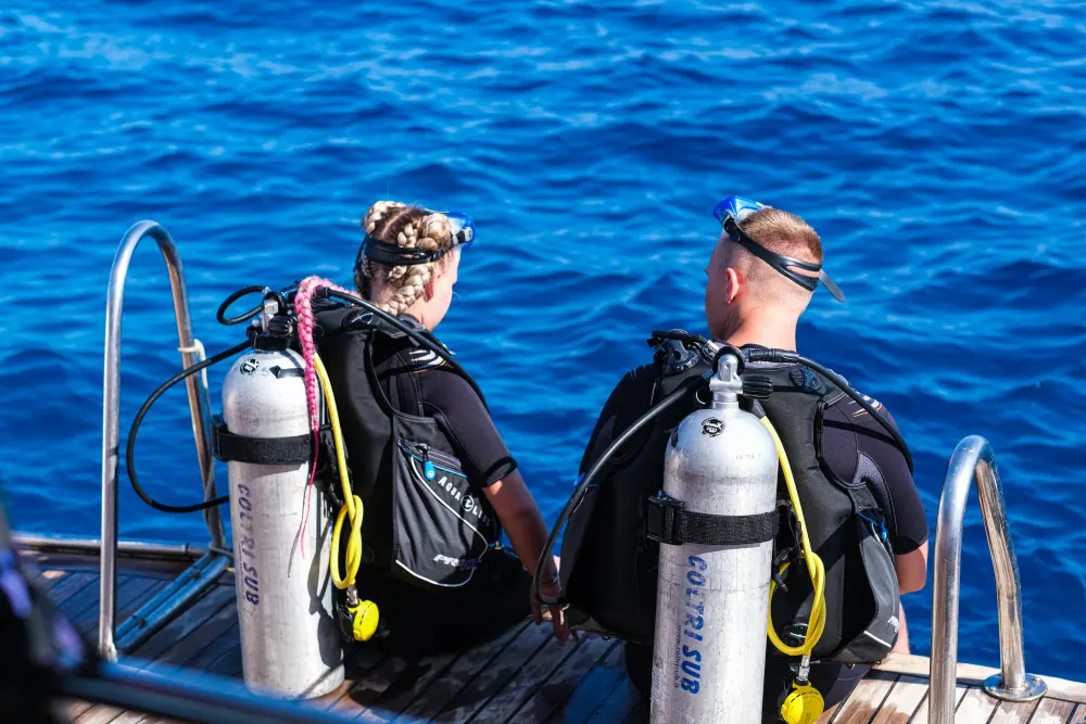 Two divers in full scuba gear sit on the side of a boat preparing to dive.