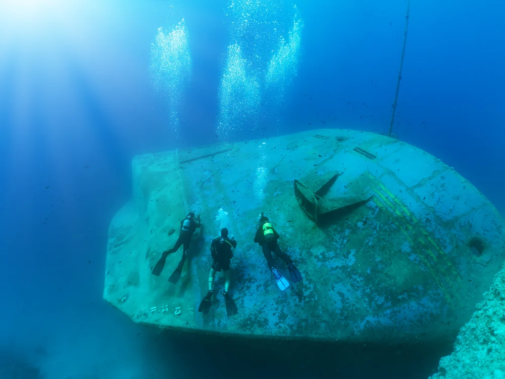 3 scuba divers swimming near the bow of a tipped over underwater shipwreck.