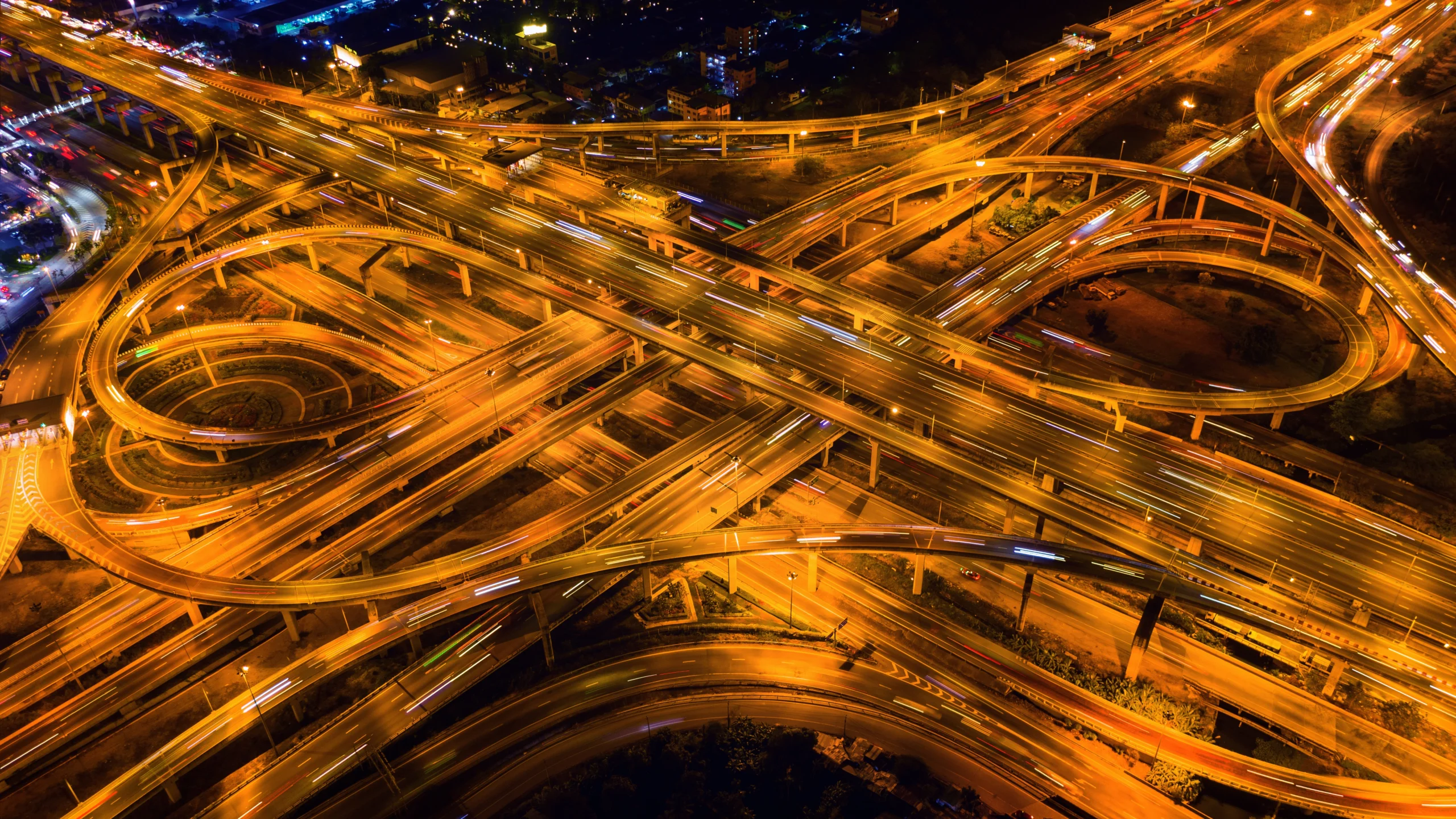 An aerial nighttime view of a complex highway interchange with multiple overpasses and roads illuminated by streetlights, showing long exposure light trails from moving vehicles.