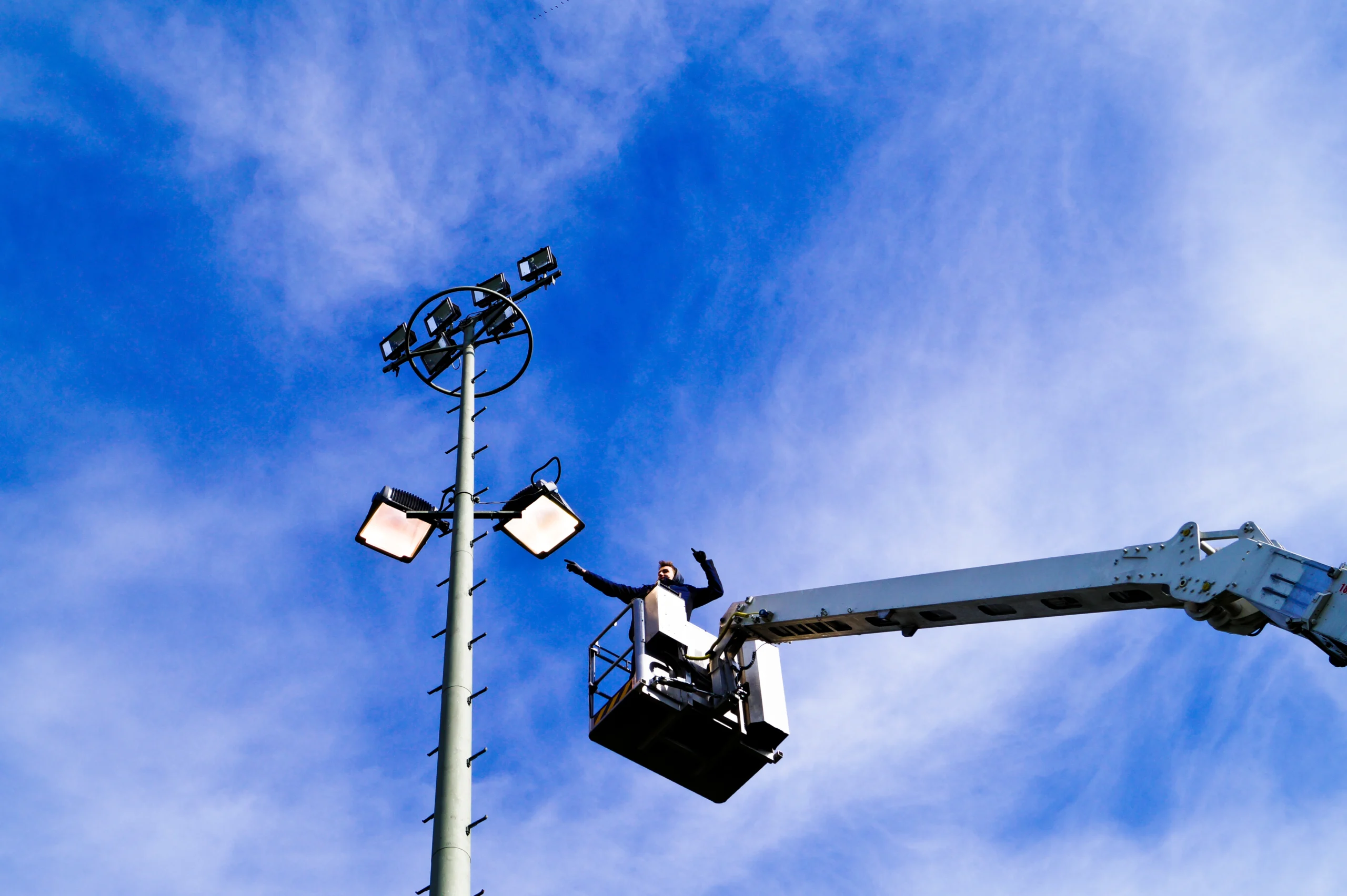 A low-angle shot shows a man in a raised bucket lift, with arms outstretched, working on a tall light pole against a blue sky with scattered clouds. The light pole has multiple spotlights and a round metal structure.