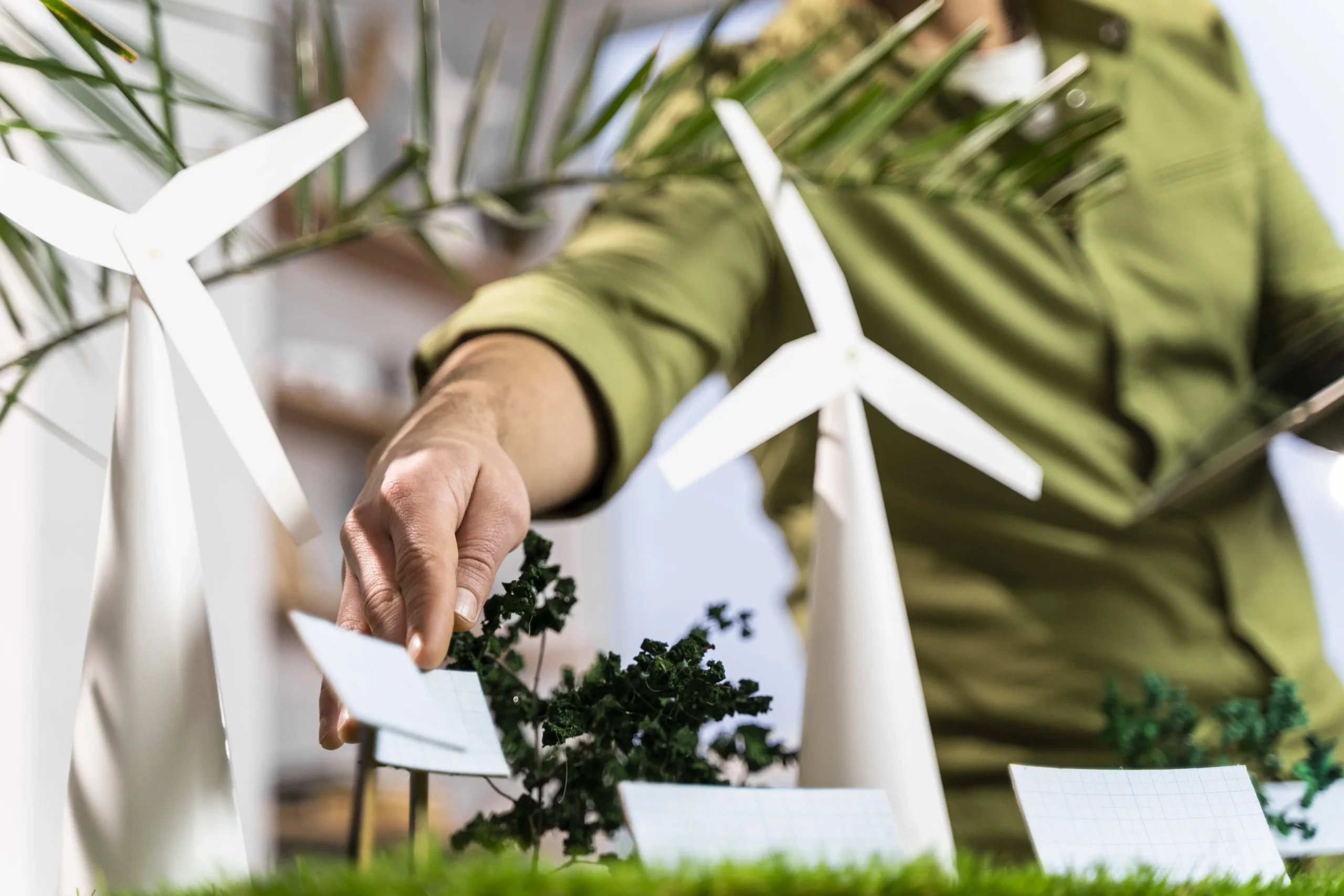 A person wearing a green shirt is arranging small, white models of wind turbines and miniature trees on a green surface, suggesting a model or diorama of a wind farm or sustainable energy project.