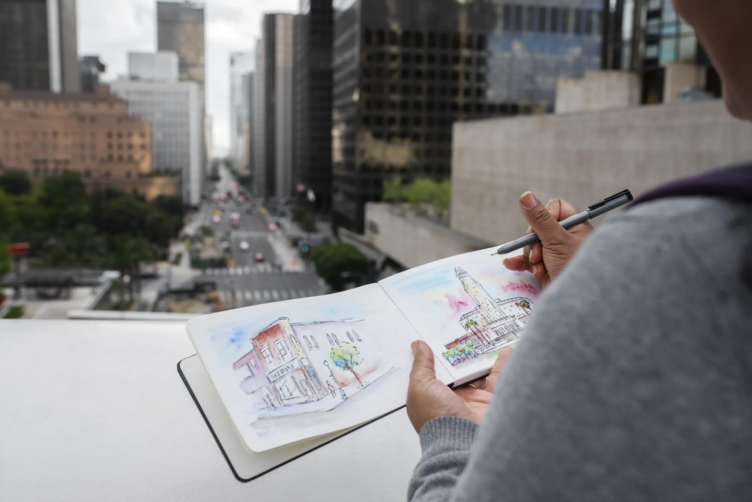 A person with their back to the viewer is sketching in a notebook, with a cityscape in the background. The notebook shows detailed line drawings of buildings, and the person is holding a pen. The background features a city street with cars and tall buildings.