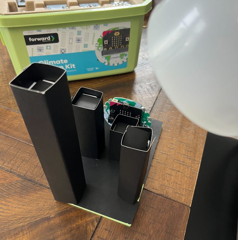 Base model city build plus dry, black construction paper coverings for Eliminating Urban Heat Islands experiment. Incandescent lamp acts as a heat/light source above the model.
