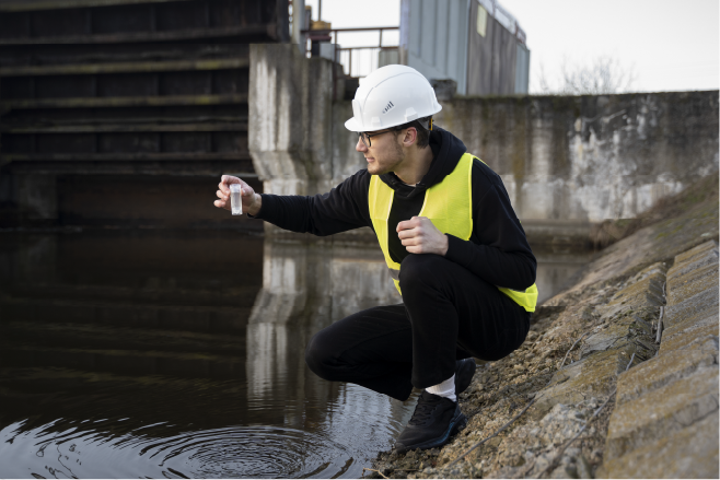 A person in a white hard hat and yellow vest crouches by a body of water, holding a small vial.