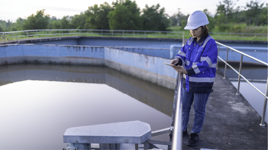 
A person in a white hard hat and blue reflective jacket stands by a large outdoor water treatment tank, looking at a tablet.