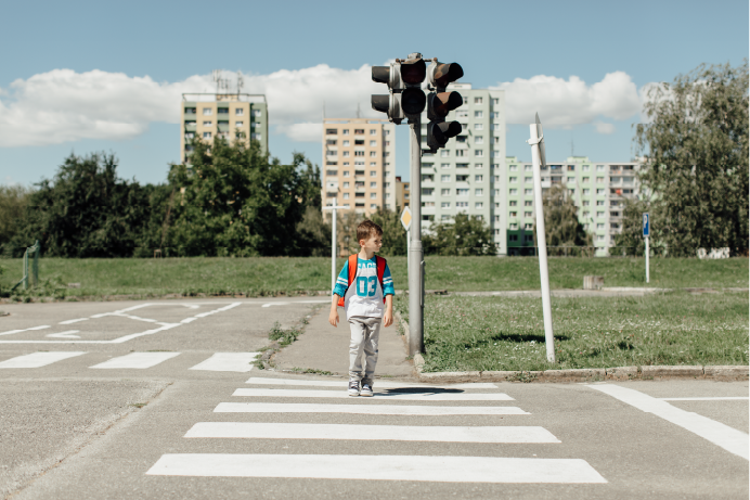 A young boy with a backpack walks across a white striped crosswalk towards a dark, non-operational traffic light on a sunny day with buildings in the background.