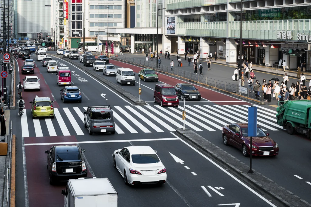 An overhead view of a bustling city street shows multiple lanes of traffic, a busy crosswalk with pedestrians, and several cars moving in both directions.