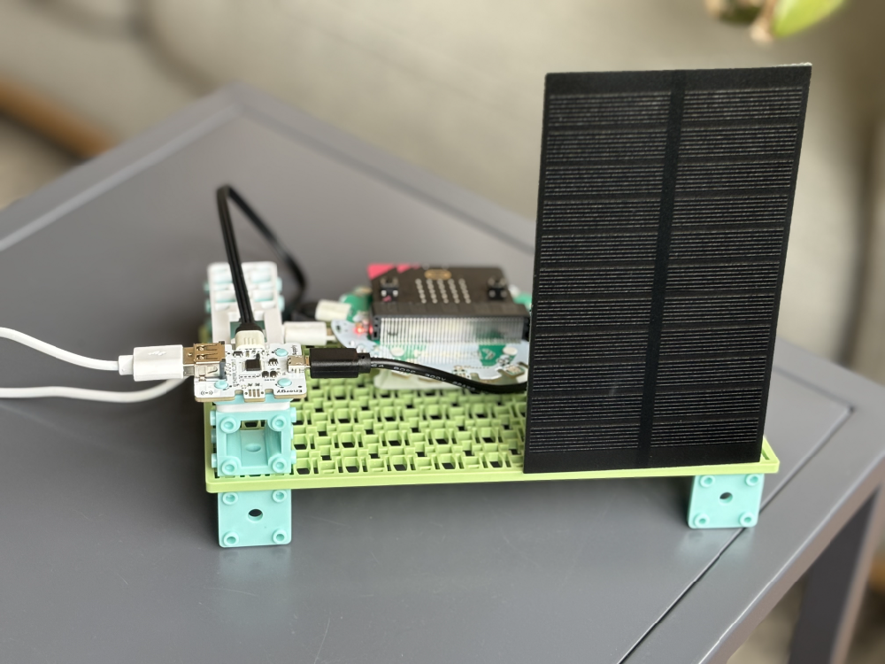 A small solar panel and circuit board assembly, featuring a micro:bit current monitor, is connected to an external device via a white USB cable.