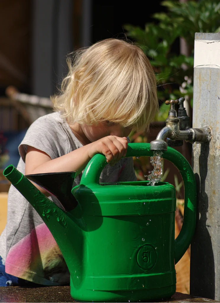 A young blonde girl refills a large green plastic watering can from an outdoor spigot.