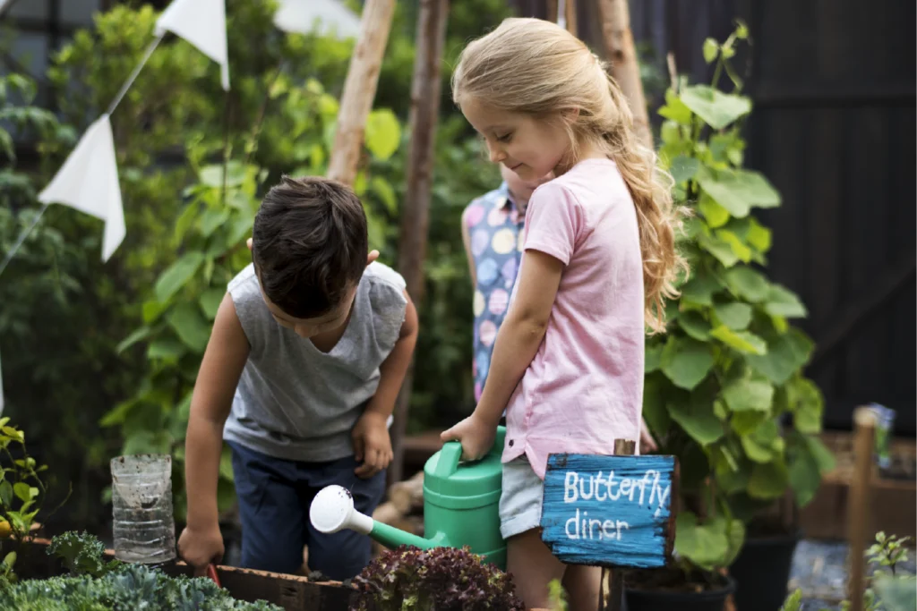 Two young children tend to plants in a garden, with the girl holding a watering can next to a blue sign that reads "Butterfly diner."