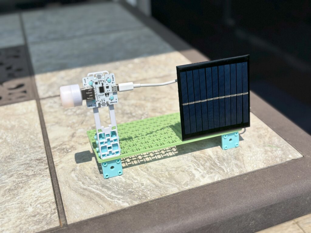 A solar-powered USB light stands on a patio table. The device includes a small, square solar panel connected by a white cord to the USB light. The components are mounted on the green, rectangular base plate included in the Smart Solar Energy Kit. The scene is lit by natural sunlight, casting shadows. 